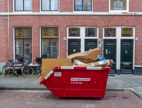 A large red industrial waste skip with a white and red safety warning label positioned on a pavement in front of a row of three dark green front doors of a brick residential building, typical of Teddington area. The skip is filled with various discarded items including brown cardboard boxes, white plastic bags, and miscellaneous debris, indicating a rubbish collection or clearance process. To the left of the skip, there are two bicycles parked near the pavement, one with a black frame and another with a dark-colored frame, secured with locks. The background features the brick facade of the building with symmetrical windows, some of which have metal grilles, and small numbered plaques next to the doors. Natural daylight illuminates the scene, highlighting the textures of the brick, metal, and rubbish contents, aligning with commercial waste removal services offered by companies like Commercial Waste Teddington in the local postcode area, serving residents and businesses in Teddington and surrounding parts of southwest London.