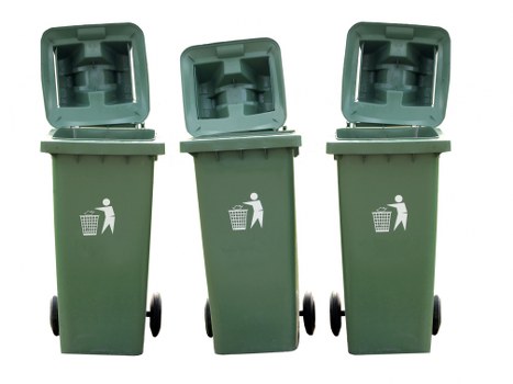 Three green wheeled rubbish bins are standing upright on a paved surface in an outdoor setting, with their hinged lids open to reveal empty interiors. The bins are made of durable plastic with a textured surface indicating typical waste container material. Each bin features a white recycling icon on the front, depicting a person disposing of waste into a bin, and is positioned in a row with slight variations in angle, suggesting they are ready for rubbish collection. The background shows a neutral outdoor environment, with no additional objects or structures visible. The scene reflects a typical waste disposal area, consistent with commercial waste management services in Teddington, and emphasizes the importance of proper waste segregation and collection. The lighting appears natural, highlighting the green color of the bins against the daylight, aligning with services provided by Commercial Waste Teddington in the local postcode area.