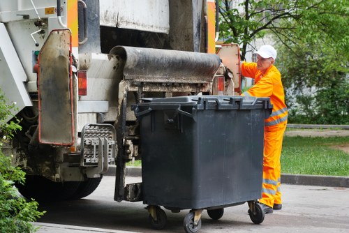 A waste collection operative dressed in high-visibility orange overalls and a white cap operates a large waste collection vehicle on a residential street. The vehicle's rear loading mechanism is engaged, with a black wheeled rubbish bin positioned beneath it, ready for collection. The bin is made of durable plastic with a textured surface, and its lid is closed. The background includes a green leafy area with trees and a sidewalk, suggesting a quiet suburban environment. The scene is well-lit with natural daylight, highlighting the practical, professional setting typical of rubbish removal services in the Teddington area, with the company's services focused on commercial waste disposal, visible from the overall context. The vehicle occupies the left side of the image, with the worker inspecting or operating controls on the right side, emphasizing the operational aspect of waste collection. The environment appears clean and orderly, fitting the context of professional waste management as provided by Commercial Waste Teddington.