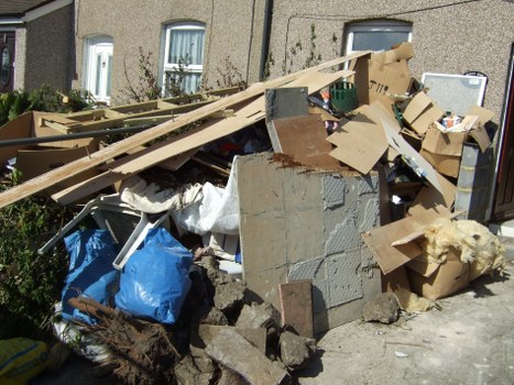 A large pile of mixed waste debris situated on a pavement outside a residential building, consisting of cardboard boxes, wooden planks, grey and brown concrete and brick fragments, and various discarded packaging materials. The cardboard boxes are partially flattened and some are torn, with a few stacked or leaning against the concrete debris. Wooden planks appear weathered, some with chipped or splintered edges, and are placed across the pile. A blue plastic bag filled with rubbish is visible among the rubble, along with white fabric or paper materials partially buried underneath. The debris extends partway up the wall of a nearby building, with windows and a small garden visible in the background. The scene is set in natural daylight, emphasizing the disorganized nature of the waste, which is likely to be removed by a professional rubbish removal service such as Commercial Waste Teddington, serving the local Teddington area.