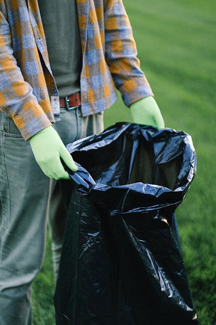 A person wearing a green long-sleeved shirt, a yellow and blue checkered jacket, light grey trousers, and light green gloves is holding open a large black rubbish bag, which is partially folded over a