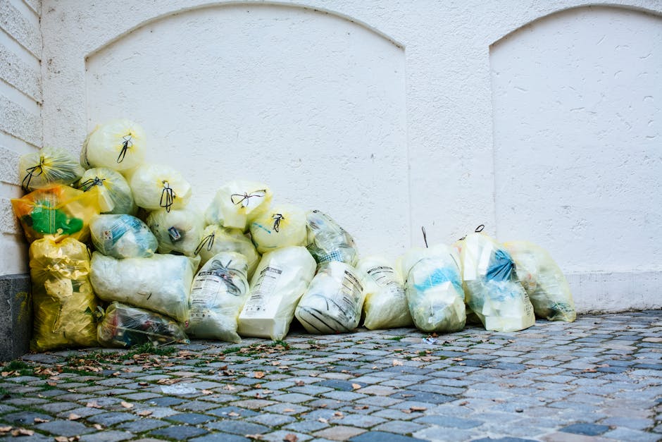 A collection of multiple refuse bags piled against a white plastered exterior wall on a cobblestone pavement. The bags are primarily yellow, white, and transparent, with some containing mixed waste or